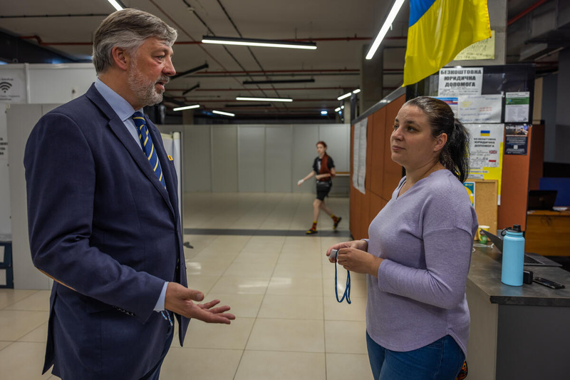 Un hombre de traje y corbata, y una mujer que viste suéter lila, conversan en una oficina.