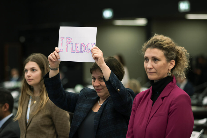 Tres mujeres forman una hilera. Las que están a los extremos son rubias; la que está en el centro, de cabello corto y oscuro, sostiene frente a sí un cartel que dice "Me comprometo".