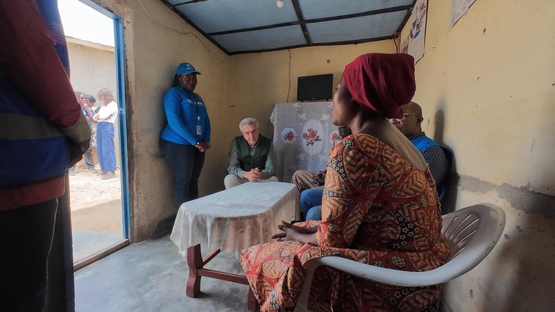 Una mujer sentada en una silla de plástico habla con personas sentadas y de pie alrededor de una mesa en el interior de un alojamiento