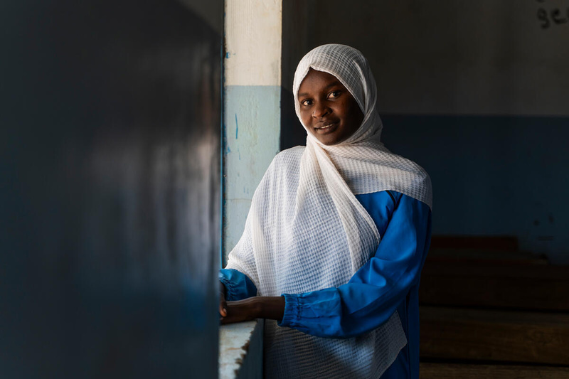 Una joven sudanesa, cuya cabeza está cubierta con un manto color blanco, sonríe a la cámara