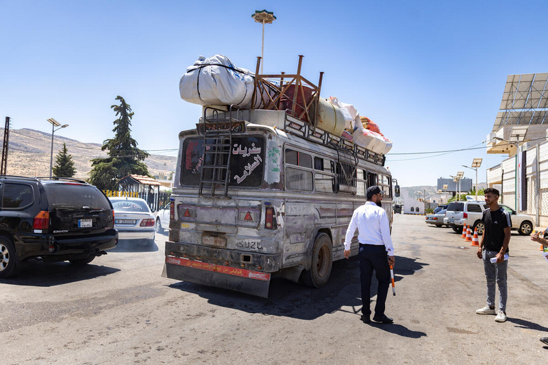 Dos hombres junto a un autobús con muebles y otras pertenencias que pasa junto a una fila de autos estacionados