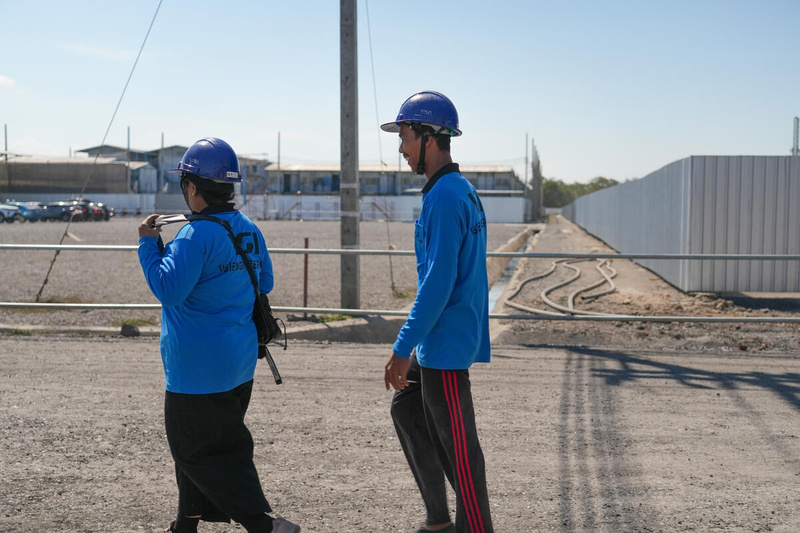 Una mujer y un hombre con uniformes azules y cascos de seguridad caminan en fila por una carretera junto a un estacionamiento