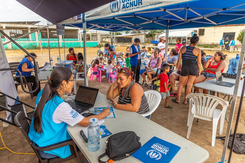 Mujeres y niños sentados en sillas bajo un gran toldo que les da sombra, acompañados por personal de ACNUR