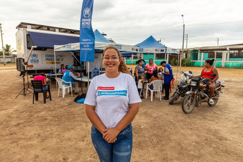Una mujer sonriente que viste camiseta blanca y pantalones de mezclilla azules está de pie frente a un grupo de personas, con una camioneta al fondo