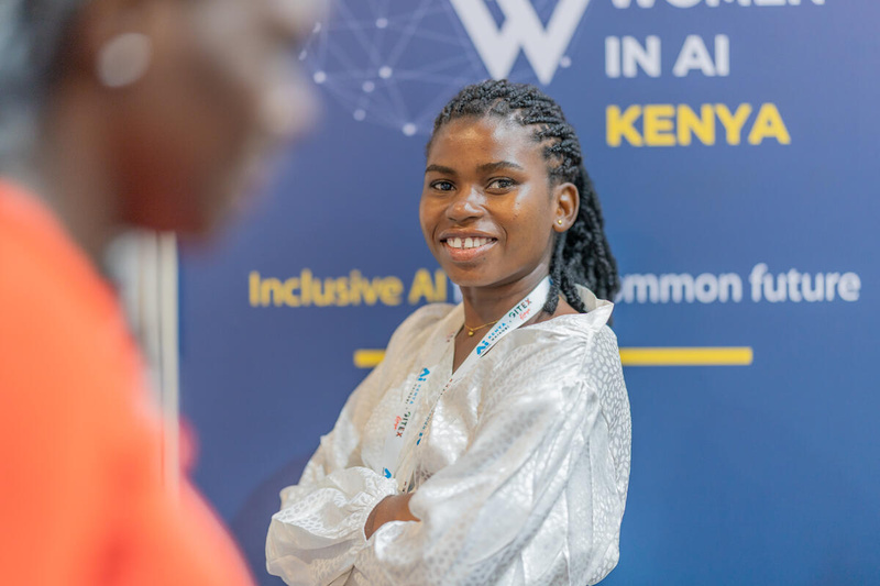 Una joven sonriente, con los brazos cruzados, posa delante del cartel de una conferencia sobre las mujeres en el ámbito de la IA en Kenia
