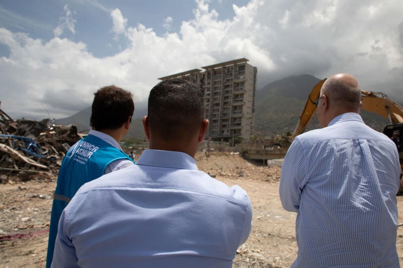 Tres hombres fotografiados de espaldas, uno de ellos con chaleco de visibilidad de ACNUR, observando un edificio de varios pisos gravemente dañado.