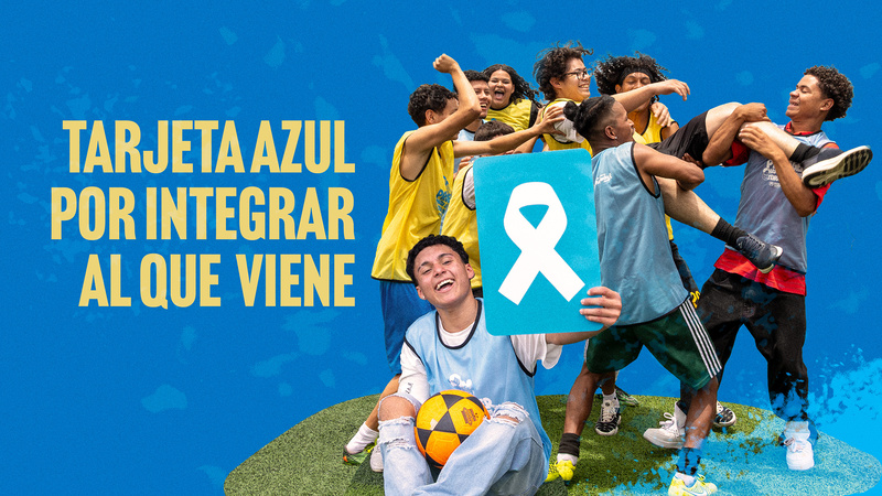 Un joven sostiene una tarjeta azul con un listón blanco en su mano izquierda y un balón amarillo con negro entre sus piernas mientras está sentado en el pasto. Al fondo, siete jóvenes cargan a un jóven