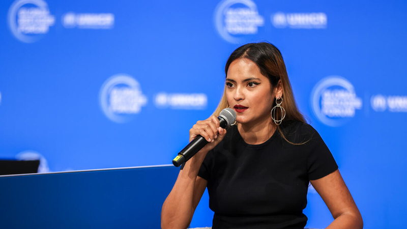 Con micrófono en mano, una refugiada rohingya pronuncia un discurso; detrás suyo, un fondo azul con un logotipo en color blanco repetido múltiples veces.