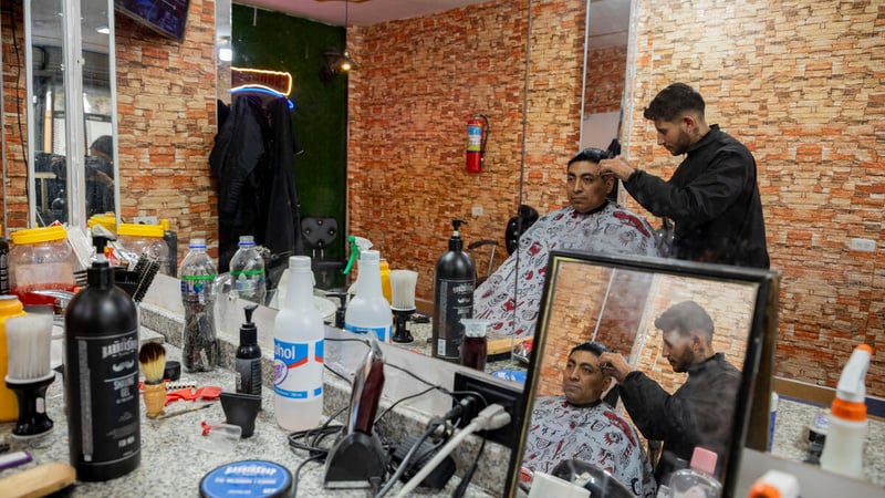 En una peluquería, un hombre joven corta el cabello de otro un poco más mayor que él.
