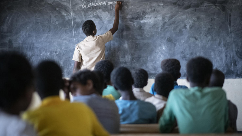 Un aula en Burkina Faso. El estudiantado le da la espalda a la cámara; el profesor escribe en el pizarrón.