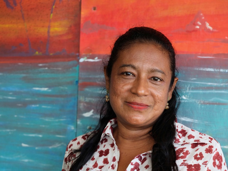 Una mujer de tez morena y cabello largo negro, que viste una blusa blanca con flores rojas, mira hacia la cámara. Al fondo, una pintura naranja con azul
