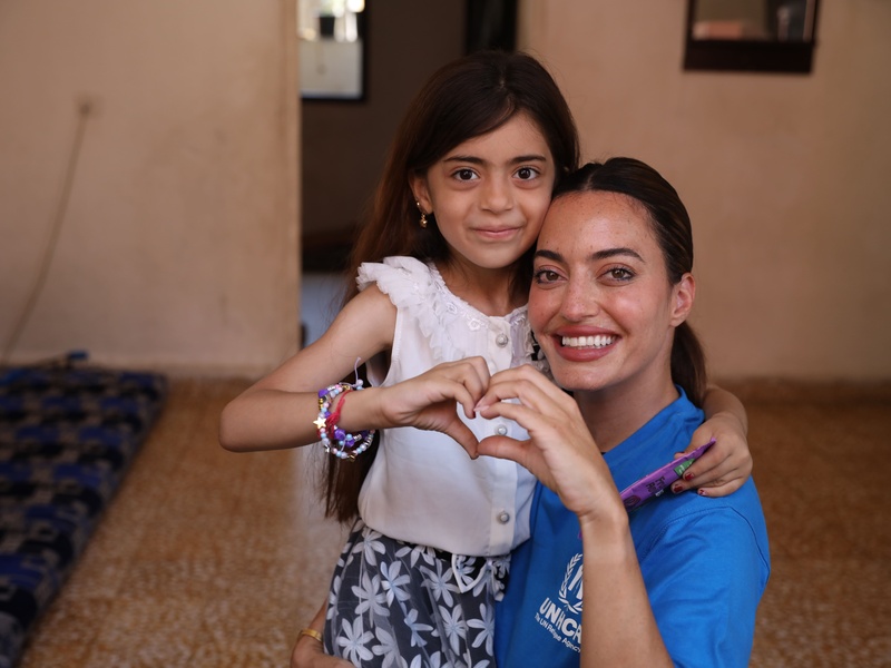 Una mujer que lleva puesta una camiseta azul con el logotipo de ACNUR abraza a una niña pequeña; con sus manos, juntas forman un corazón.