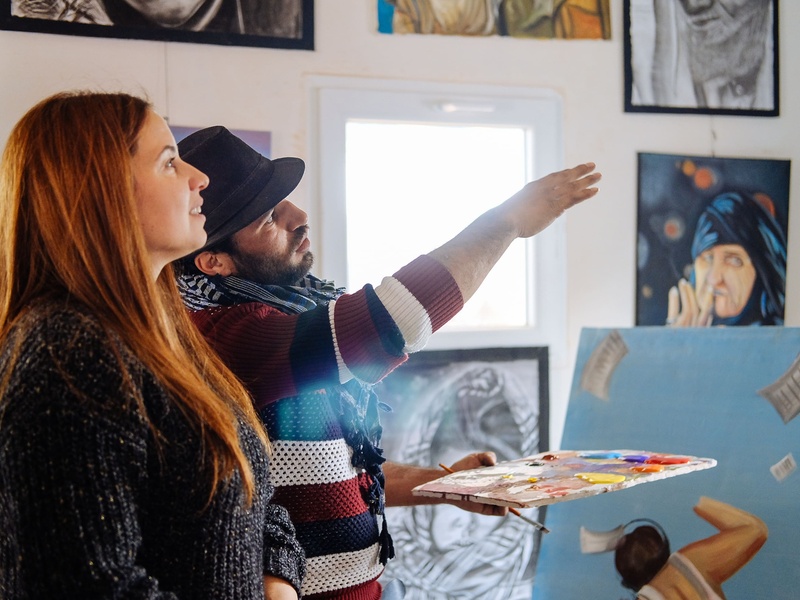 En un estudio de arte, una mujer escucha a un hombre mientras este explica sus pinturas.