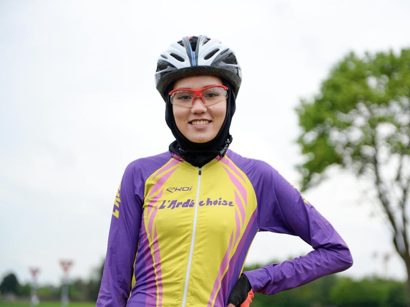 Una joven ciclista sonríe a la cámara; lleva puestas gafas y un hijab debajo del casco. Detrás suyo hay un árbol.