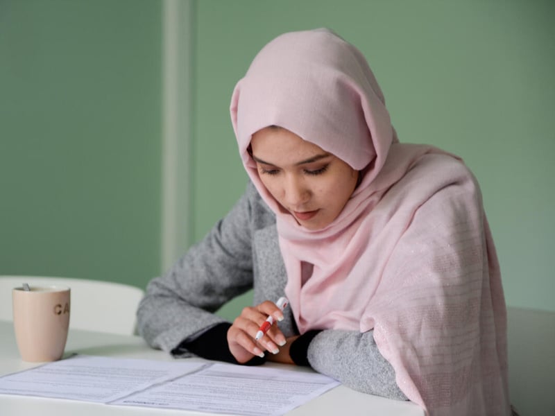 Una joven que lleva puesto un hijab color durazno está estudiando en un escritorio; junto a los papeles que tiene frente a sí hay un vaso.