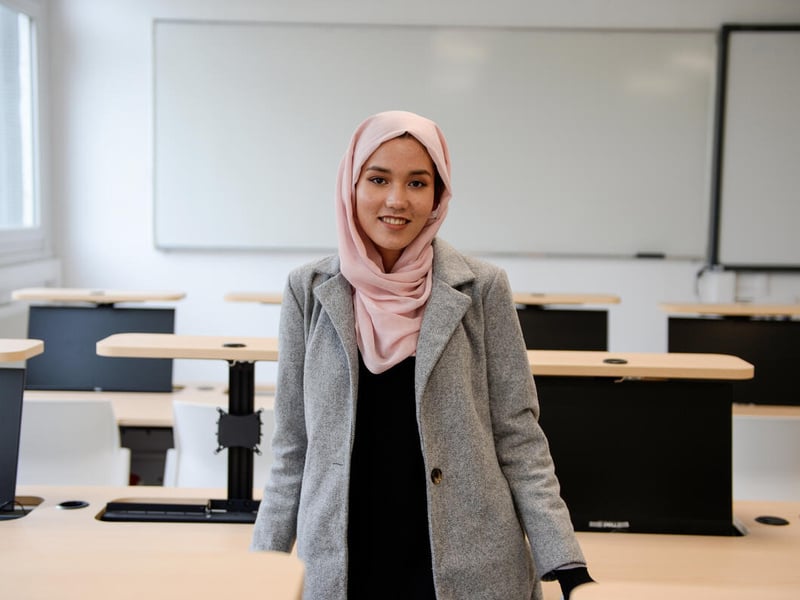 Una joven de Afganistán sonríe a la cámara desde el centro de un aula. Lleva puesto un hijab color durazno; detrás suyo, un pintarrón.