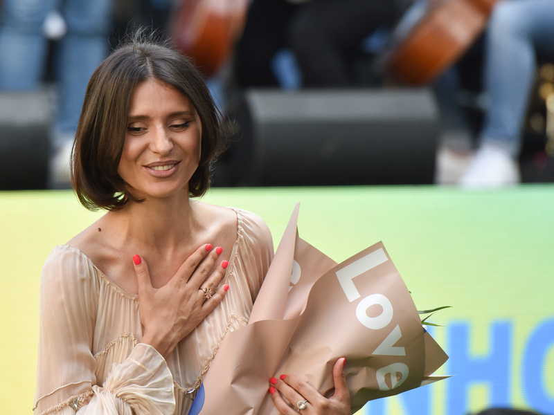 Una joven de cabello castaño que lleva puesto un vestido color beige sonríe sosteniendo un ramo de flores