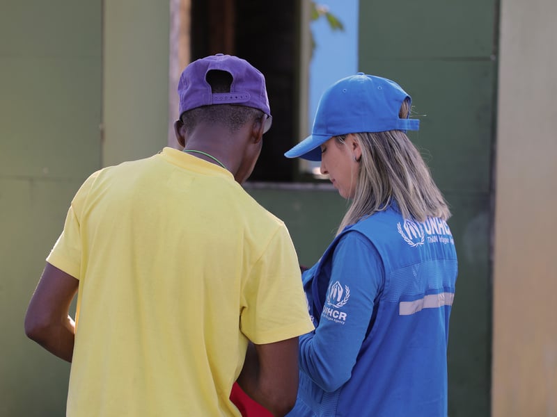 De espaldas un hombre de gorra morada y camiseta amarilla, junto a una mujer de cabello rubio que porta gorra y chaleco azul de ACNUR
