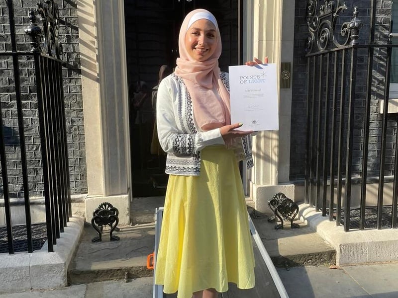 Una joven siria, que lleva puesto un hiyab, sonríe sobre una acera mientras muestra un documento a la cámara.