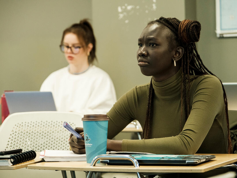 Dos jóvenes, una caucásica y una negra, en un aula universitaria. En primer plano, la joven negra, originaria de Sudán del Sur, mira al frente del aula con atención.