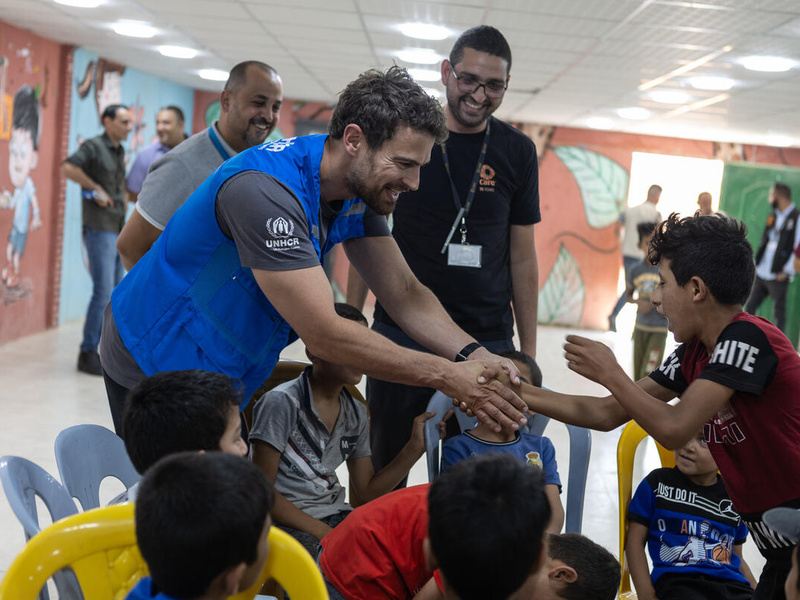 Un hombre joven que lleva puesto un chaleco con el logotipo de ACNUR salud a un grupo de niños en un campamento de refugiados en Jordania