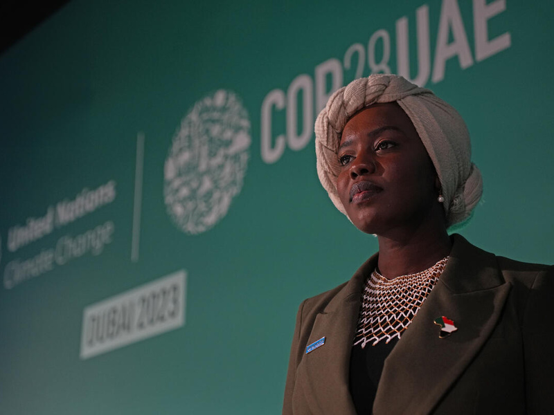 Una mujer sudanesa-estadunidense mira al horizonte; detrás suyo, una pancarta verde que dice COP28 en letras blancas.