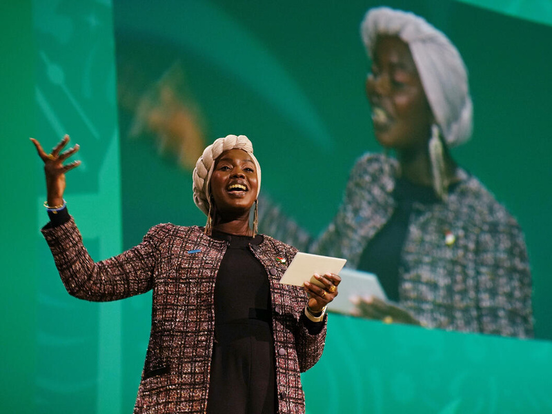 Una mujer sudanesa-estadunidense, que lleva puesta una especie de turbante color blanco, recita apasionadamente un poema; detrás suyo, una pantalla verde en la que se proyecta una imagen de ella misma.