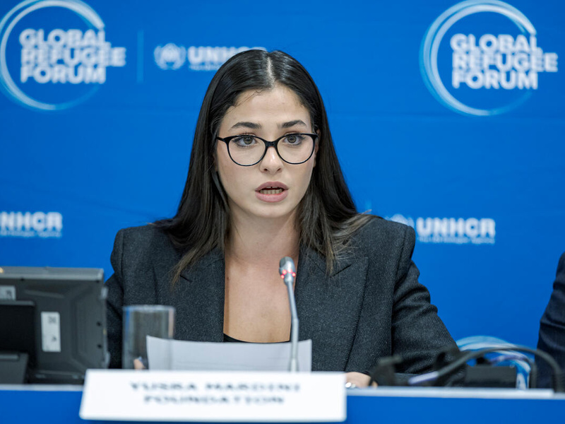 Una mujer joven, que usa anteojos, pronuncia unas palabras frente a un micrófono; detrás suyo, un fondo azul con el logotipo del Foro Mundial sobre los Refugiados.