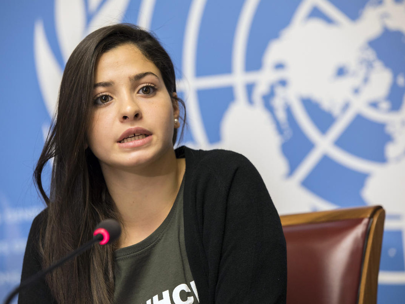 Una mujer joven que parece estar hablando; detrás suyo, un fondo azul en el que se observa una parte del logotipo de las Naciones Unidas.
