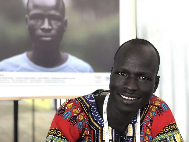 Un hombre sursudanés, que lleva puesta una colorida indumentaria, sonría a la cámara; detrás suyo, una fotografía de él mismo.