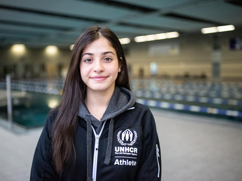 Una mujer joven que lleva puesta una chaqueta negra con el logotipo de ACNUR sonríe a la cámara; detrás suyo, una piscina.