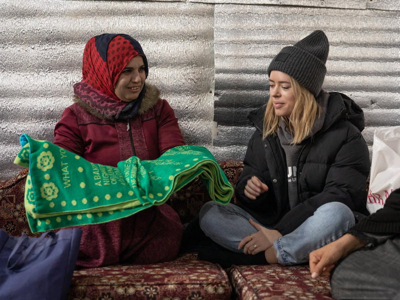 Una mujer que lleva puesto un jiyab le muestra una manta verde con decoración amarilla a una mujer caucásica que lleva puesto un gorro de estambre