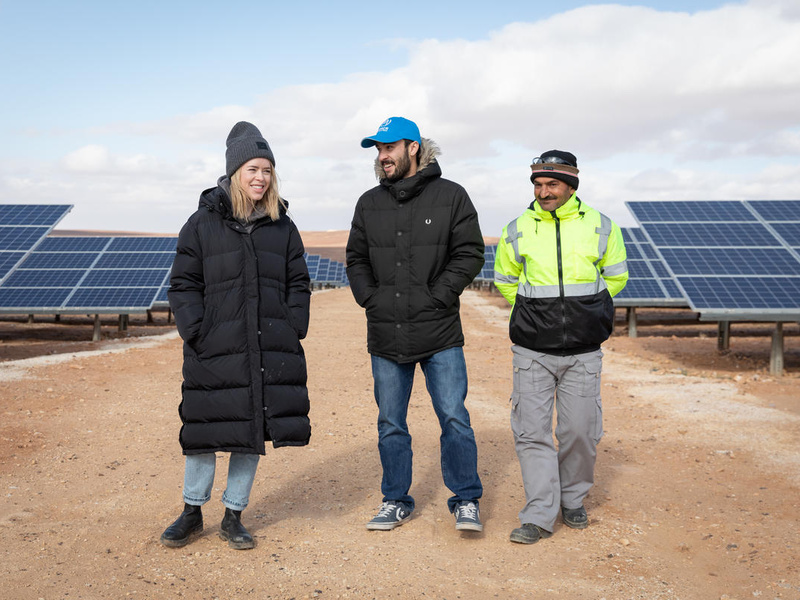 Una mujer caucásica con una abrigo negro largo charla con dos hombres en medio de un área con paneles solares