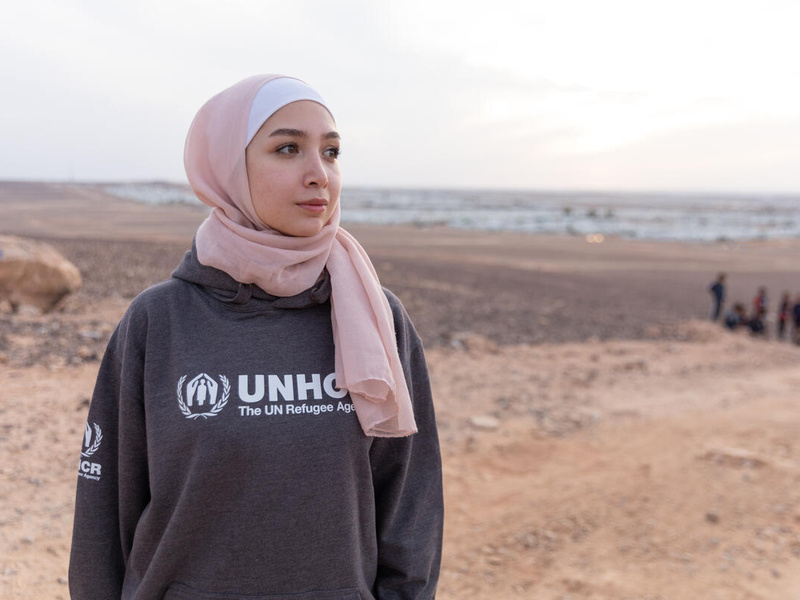 Una joven siria, que lleva puesto un hiyab rosado, mira al horizonte en medio de un terreno vacío; al fondo, un campamento de refugiados.