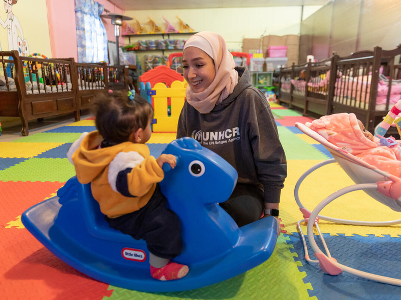 Una joven siria, que lleva puesto un hiyab rosado, le sonríe a una pequeña niña que está montada en un juguete azul con forma de caballo en un espacio de juegos infantiles.
