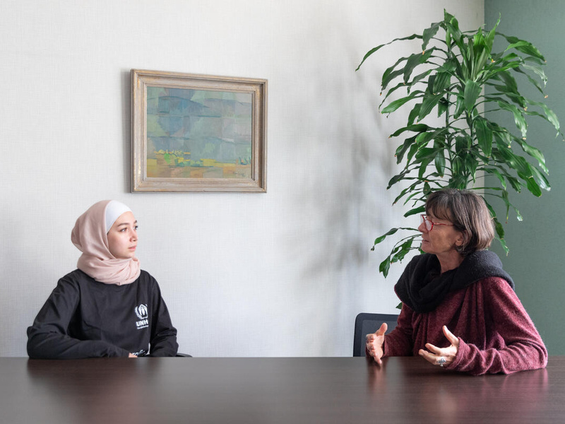 Dos mujeres, una de ellas (la que está del lado izquierdo) lleva puesto un hiyab, charlan sentadas frente a una mesa de madera.