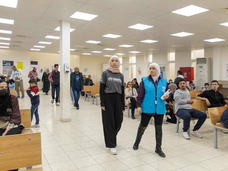 Dos mujeres, ambas llevan puesto un hiyab, recorren el pasillo de un centro de registro; a los lados, bancas ocupadas por personas que esperan.