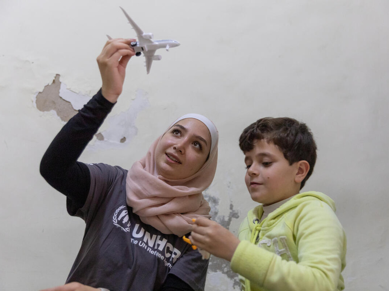 Una joven siria, que lleva puesto un hiyab rosado y sostiene un avión de juguete con la mano derecha, junto a un niño sirio que lleva puesta una sudadera amarilla.