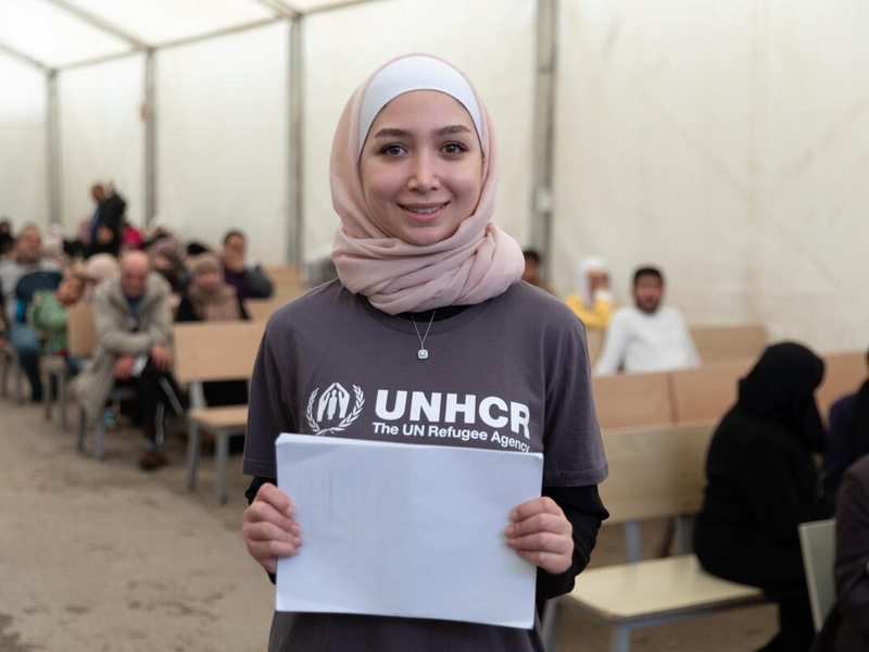 Una joven siria, que lleva puesto un hiyab rosado y sostiene unos papeles frente a sí con ambas manos, sonríe a la cámara en medio de un pasillo; detrás suyo, varias personas sentadas en bancas de madera.