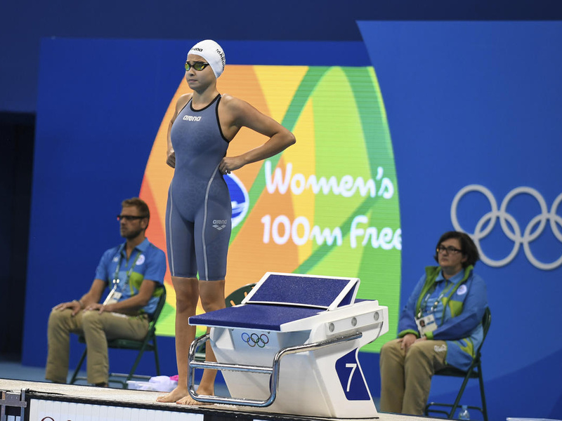 Una nadadora junto a la plataforma de salida en una piscina de los Juegos Olímpicos de Río de Janeiro; detrás suyo, un fondo azul y dos personas (un hombre y una mujer) sentadas en sillas plegables.