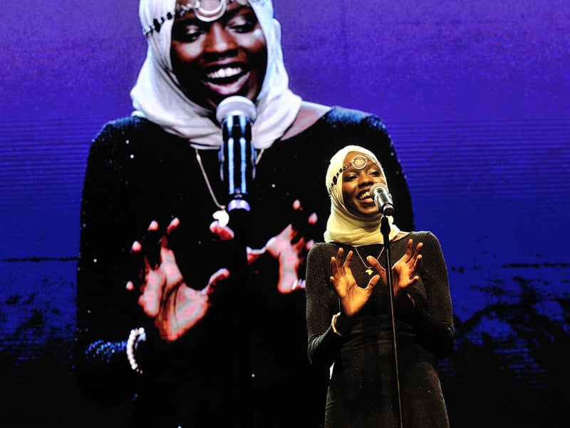 Una poeta sudanesa-estadunidense, que lleva puesto un hiyab, recita un texto delante de un micrófono; detrás suyo, una pantalla en la que se proyecta una imagen de ella de gran tamaño.