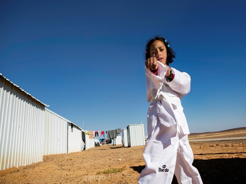 En un campamento de refugiados, una niña que lleva puesto un uniforme blanco de karateka hace un movimiento de karate frente a la cámara