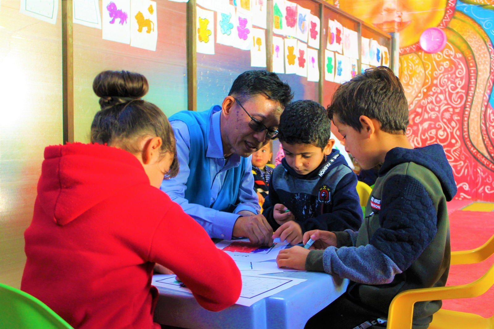 El Sr. Wanchai Tachavejnukul dibujando junto a niñas y niños, sentado a una mesa en en aula escolar.