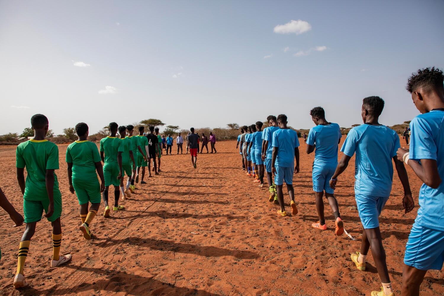 Two lines of players wearing green and blue kits walk onto a dusty soccer pitch.