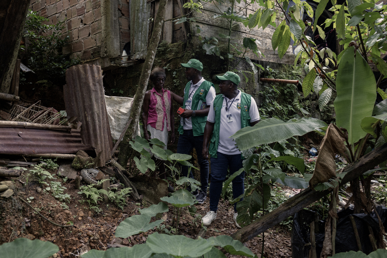 Dos voluntarios con chalecos verdes visitan a una mujer mayor de aspecto fr&aacute;gil en su choza, en una zona apartada de la selva.