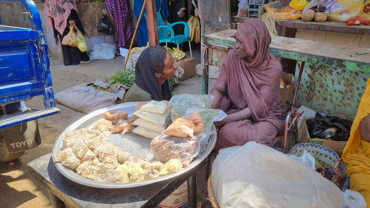 Una mujer se agacha para hablar con otra sentada frente a una bandeja de comida que vende en un mercado.