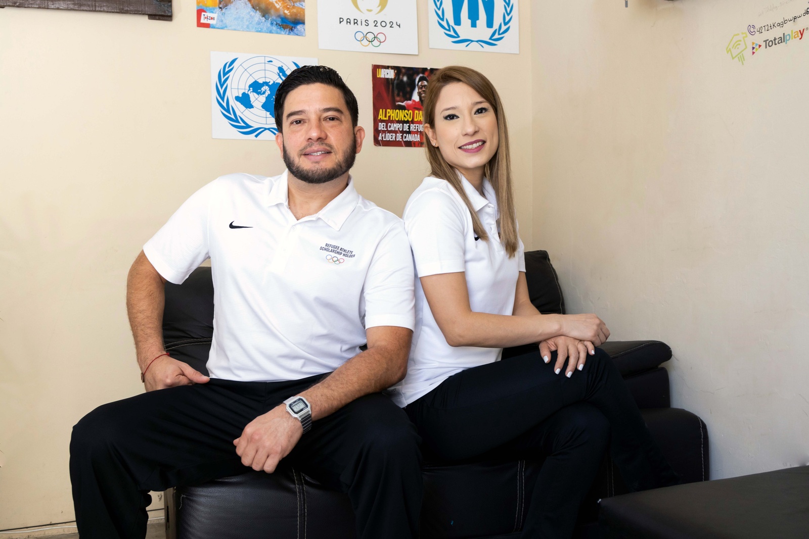 Un hombre y una mujer visten playera blanca y pantal&oacute;n negro. Al fondo, carteles del logo de ACNUR y de las Olimpiadas 2024