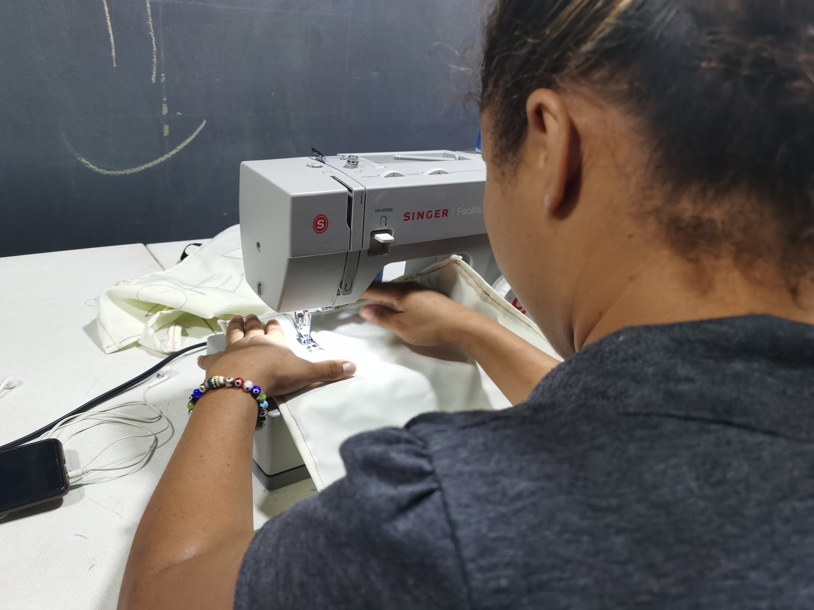 Foto de espaldas a una mujer con camiseta gris oscuro que utiliza una m&aacute;quina de coser