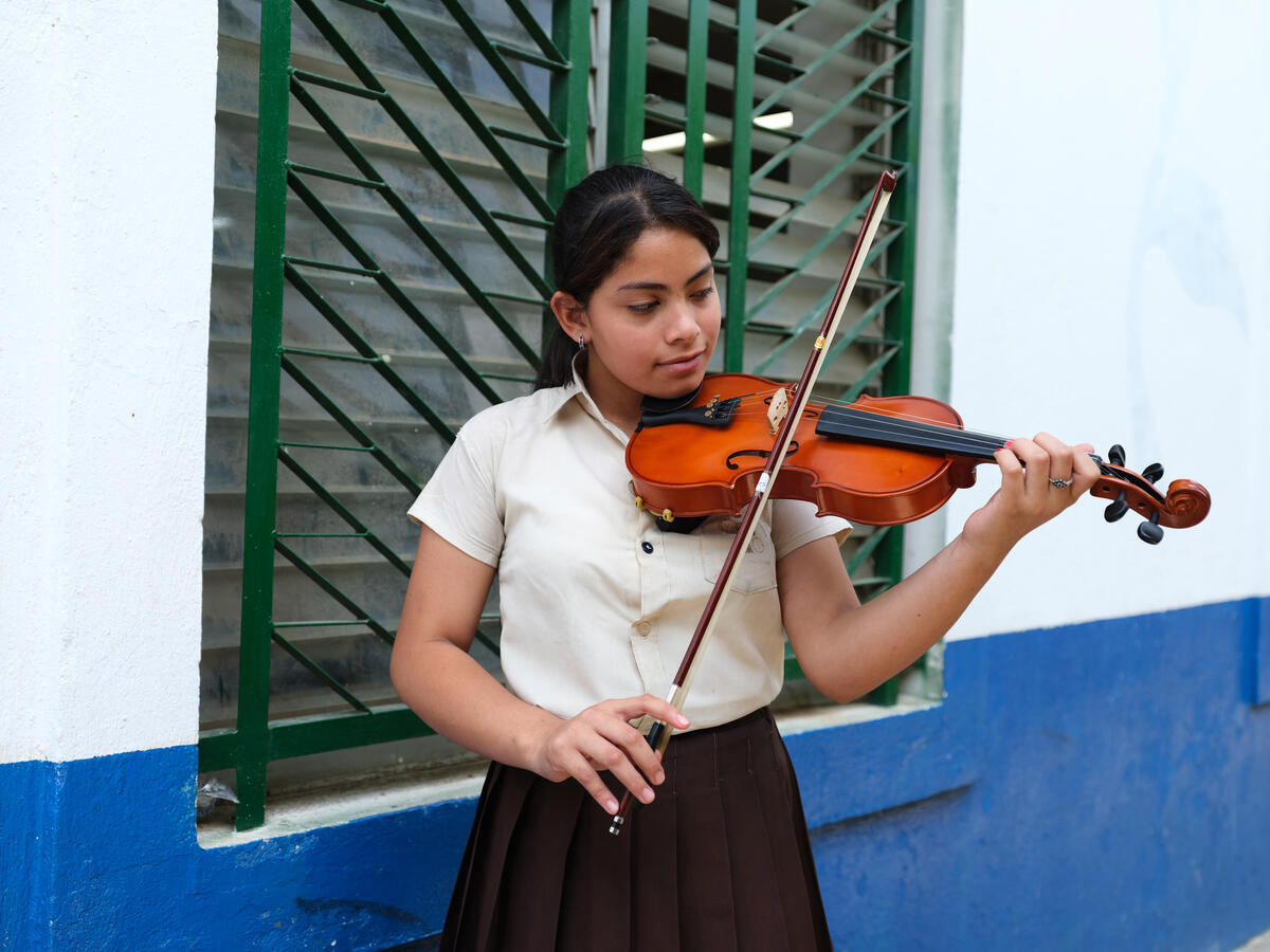 Una niña con uniforme escolar toca el violín frente a una ventana