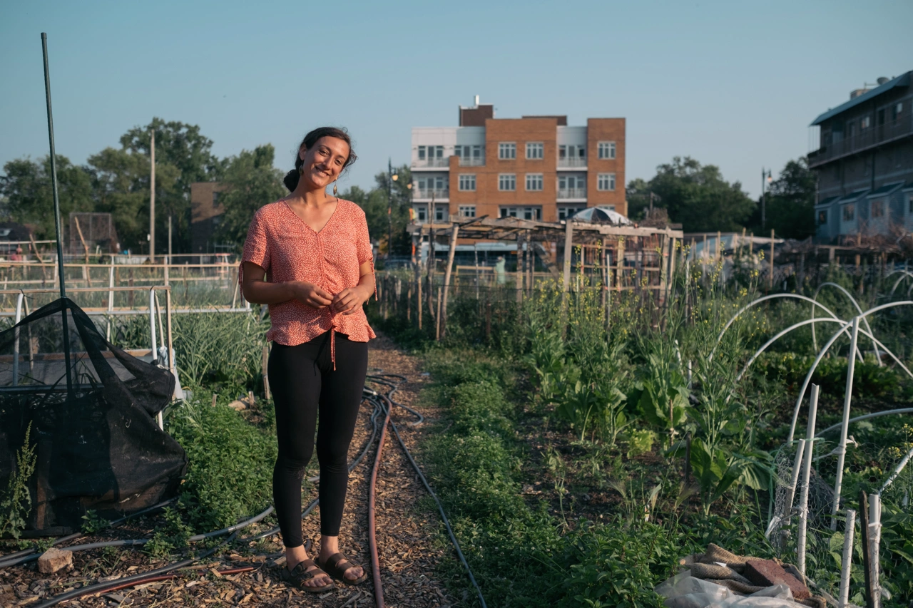 Una mujer joven sonríe a la cámara; está de pie en medio de un lote lleno de vegetación.